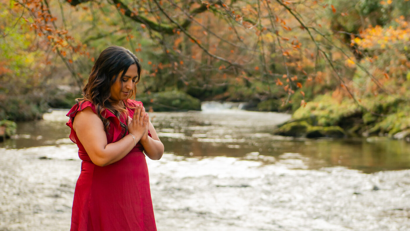 Photograph of Helen Biswas with her hands in a prayer position standing near a river