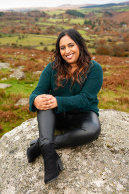 Photograph of Helen Biswas sittingon a rock with country side behind her.